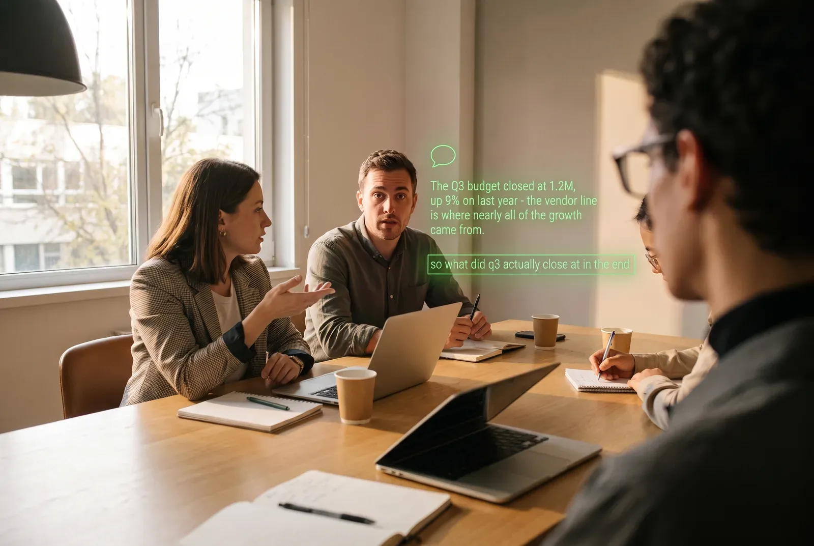 Seen through the glasses: a meeting room, three colleagues across the table. On the lens: a speech-bubble icon, then: The Q3 budget closed at 1.2M, up 9% on last year - the vendor line is where nearly all of the growth came from. Below it: what was just said.